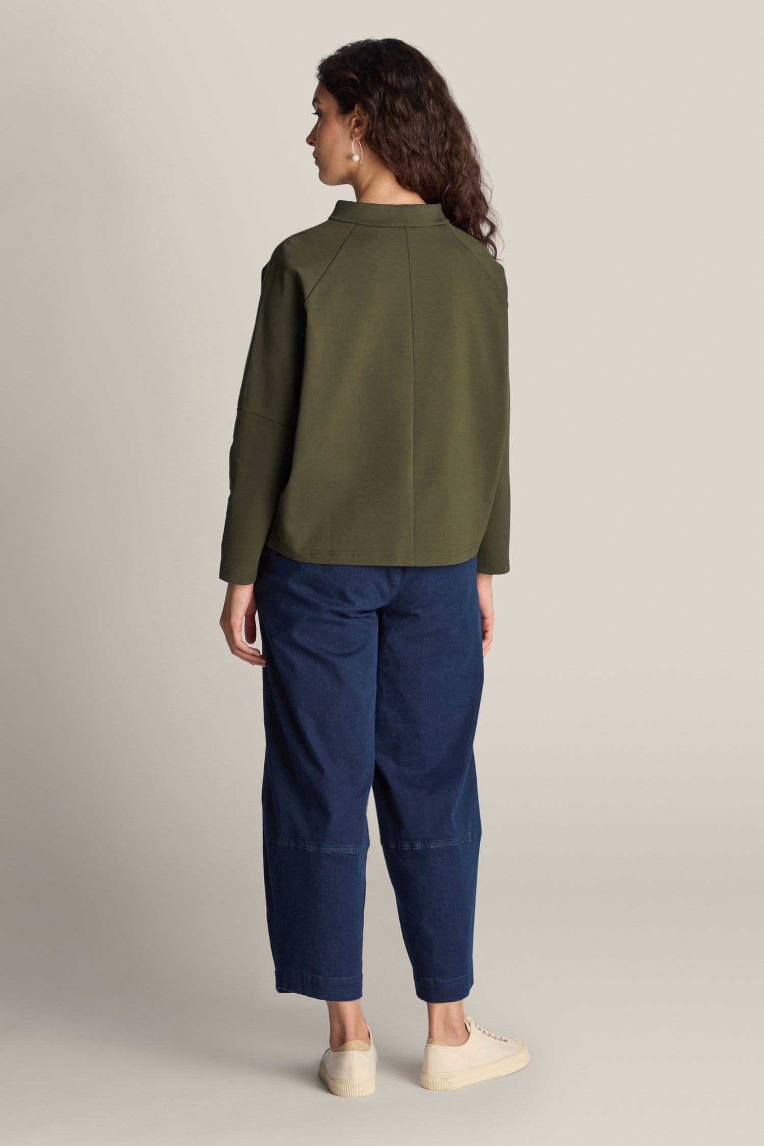 A person with long, curly hair is standing with their back to the camera, wearing a chic green Ponte Boxy Top. This top enhances their casual look, which they have paired with blue pants and white shoes.