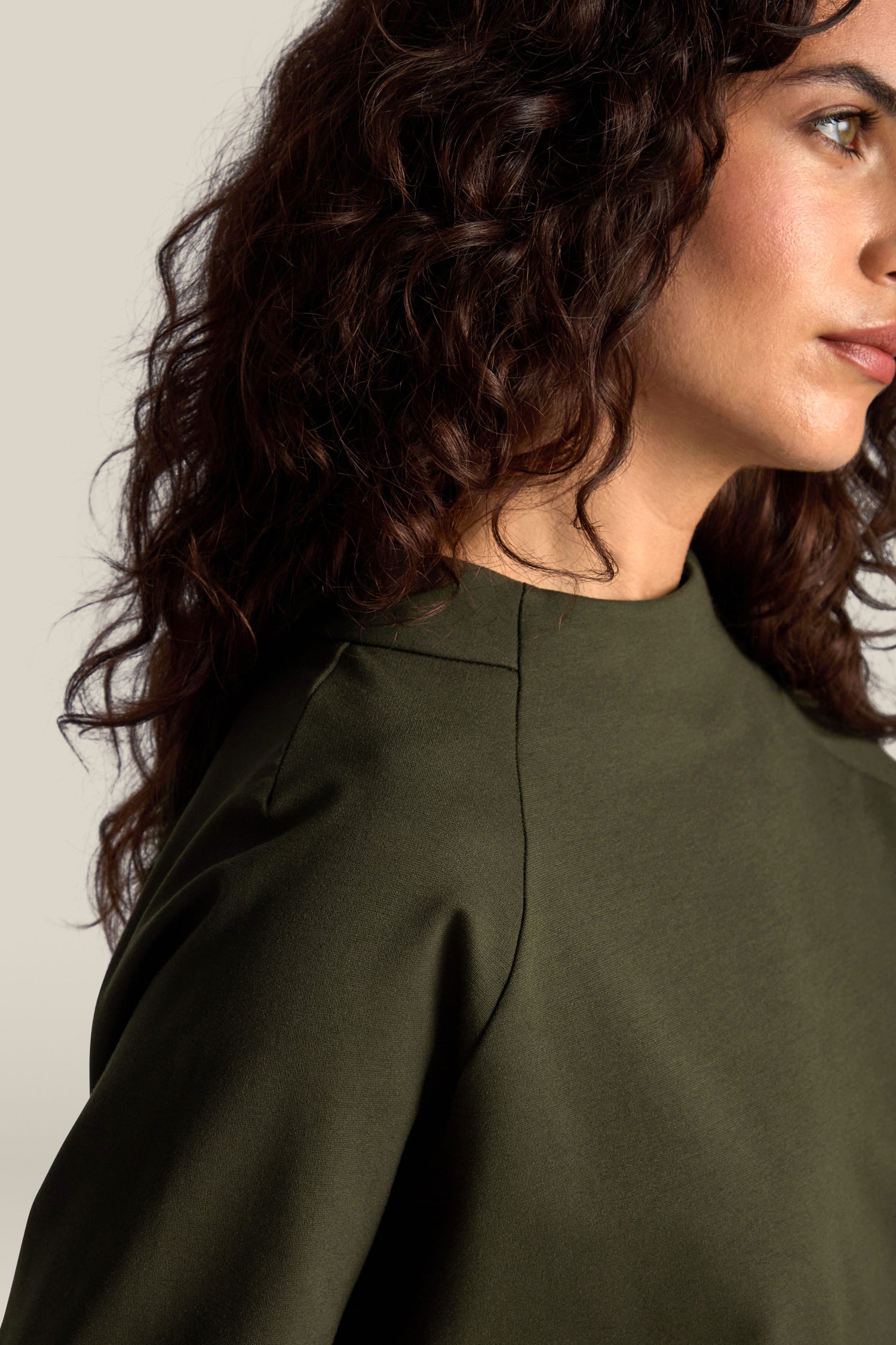 A close-up of a person with long, curly hair wearing the Ponte Boxy Top in dark green against a light gray background, showcasing a casual chic look.