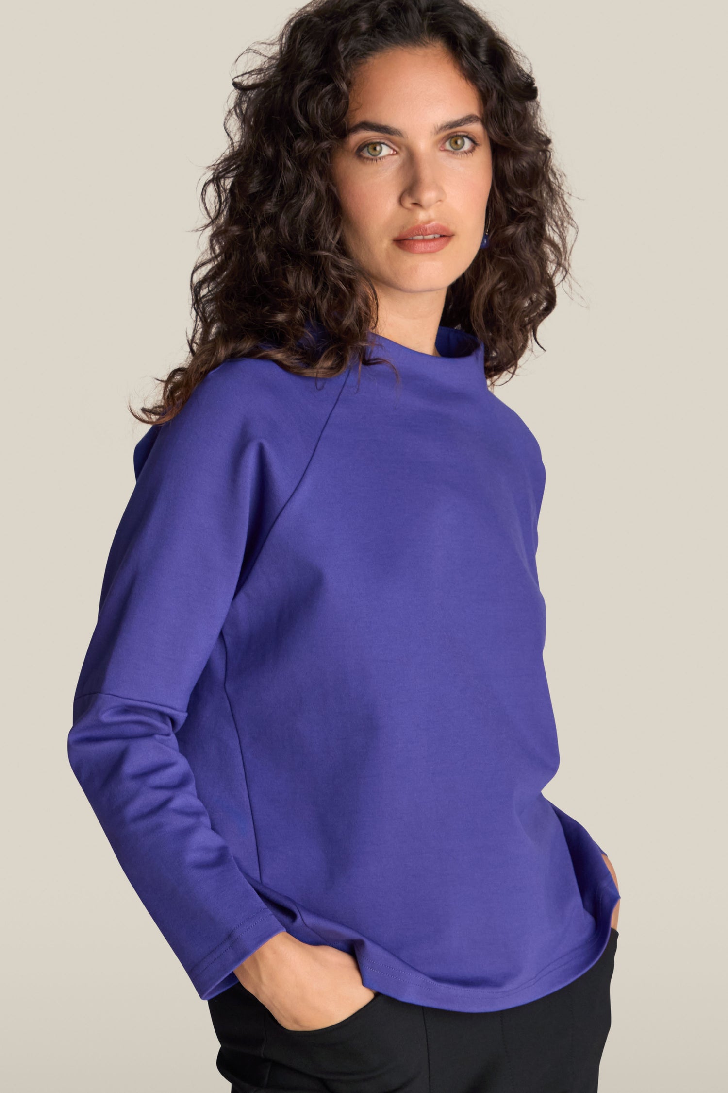 A woman with curly brown hair is wearing the Ponte Boxy Top, a blue long-sleeve high-neck top, paired with black pants. Her hands are in her pockets as she stands against a plain background. The boxy design of the top adds a contemporary edge to her casual chic look.