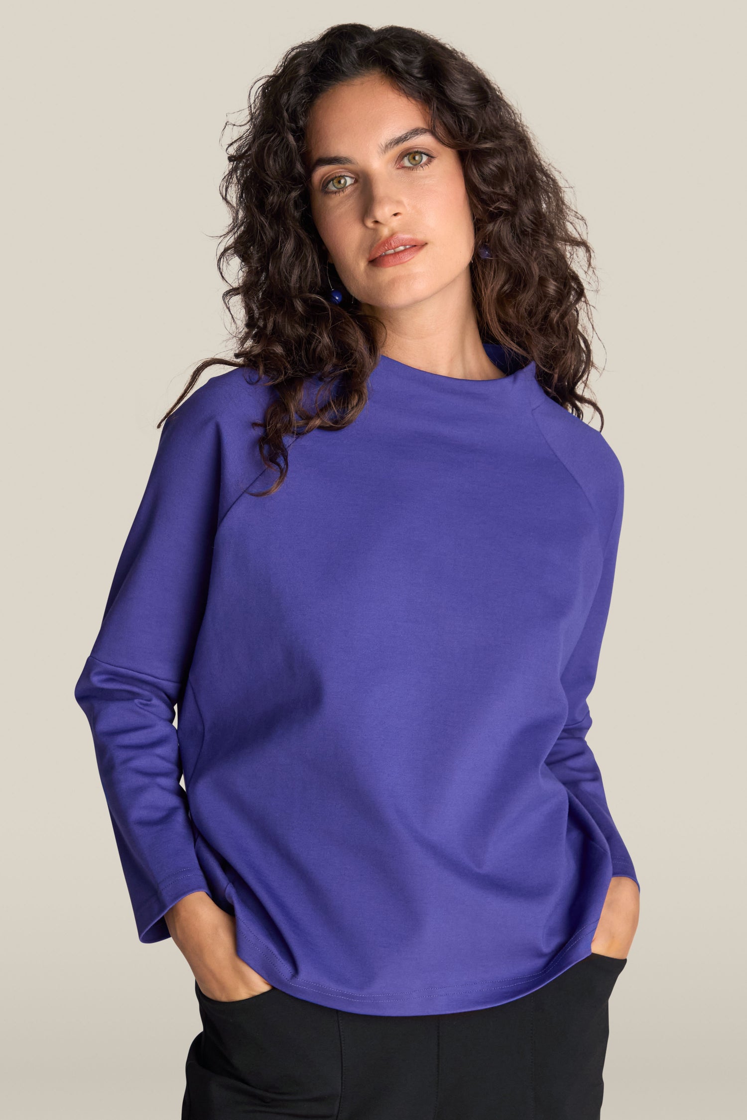 A person with long curly hair wearing a purple Ponte Boxy Top and black pants stands with their hands in their pockets against a plain background, showcasing a casual chic look.