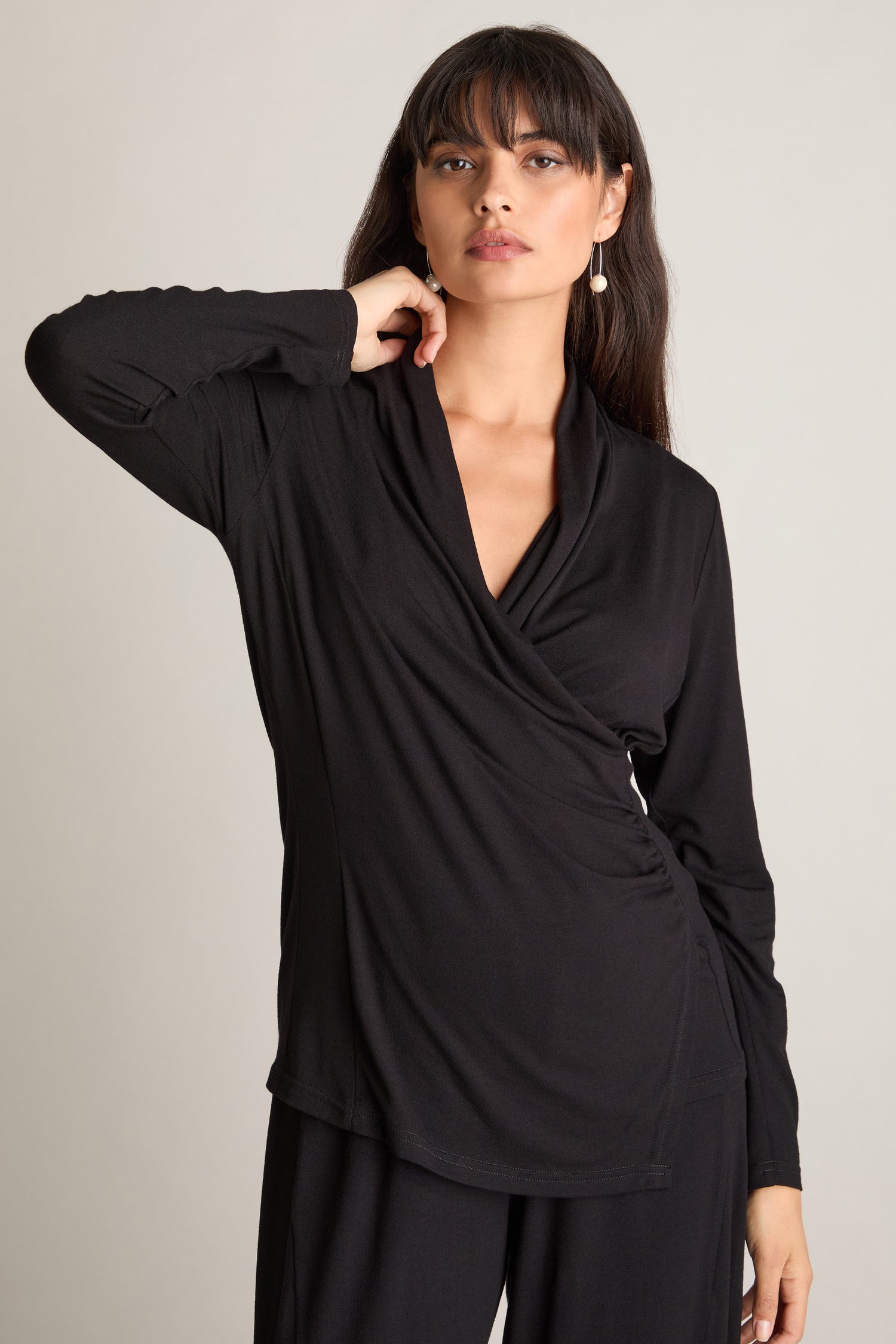 A woman wearing a Fluid Crepe Jersey Ruched Side Top stands against a neutral background, touching her chin with one hand.