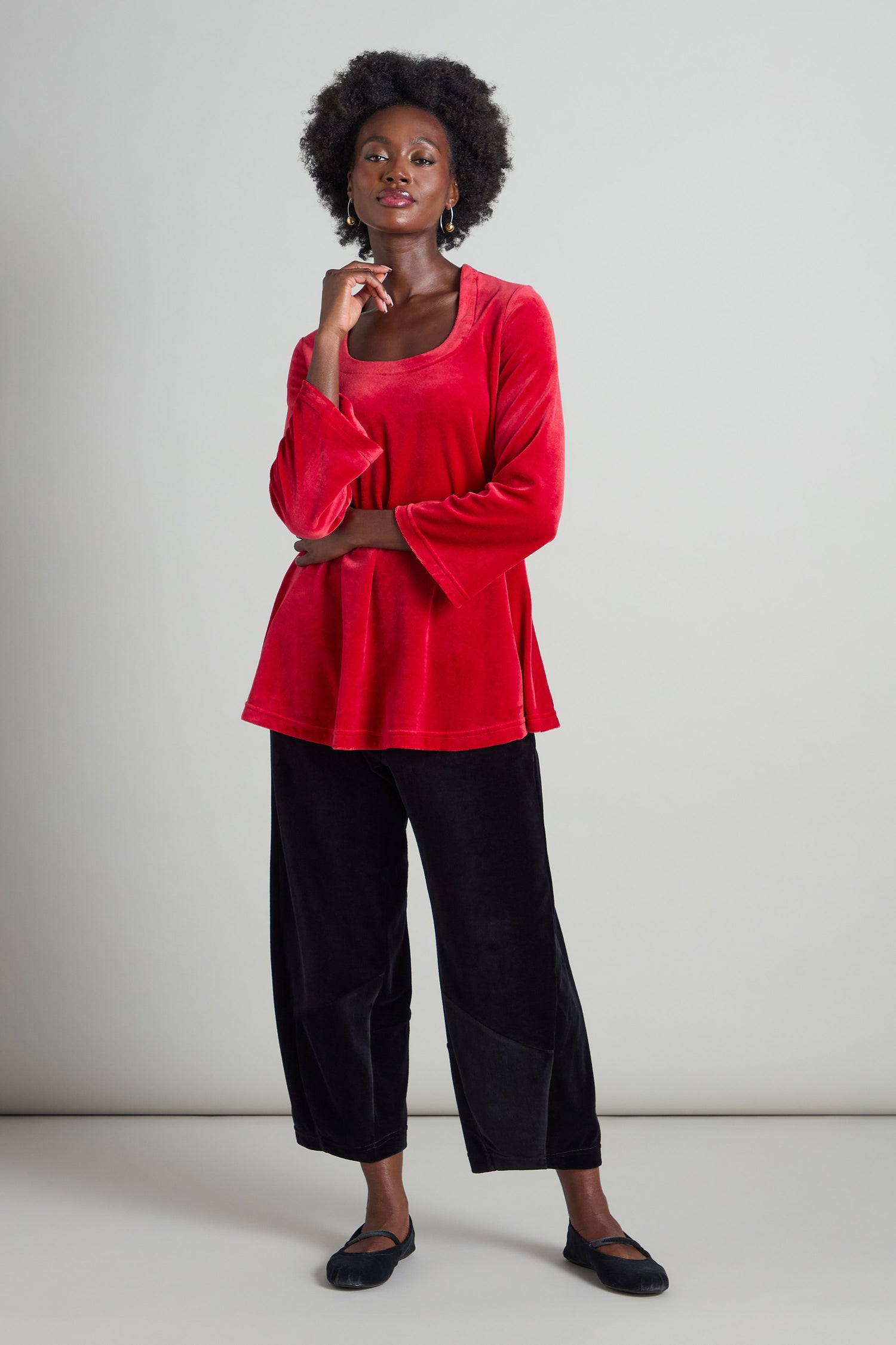 A person wearing the Velvet Jersey Horseshoe Neck Top in red, paired with black velvet pants, stands against a plain background with one hand on their chin and the other arm crossed.