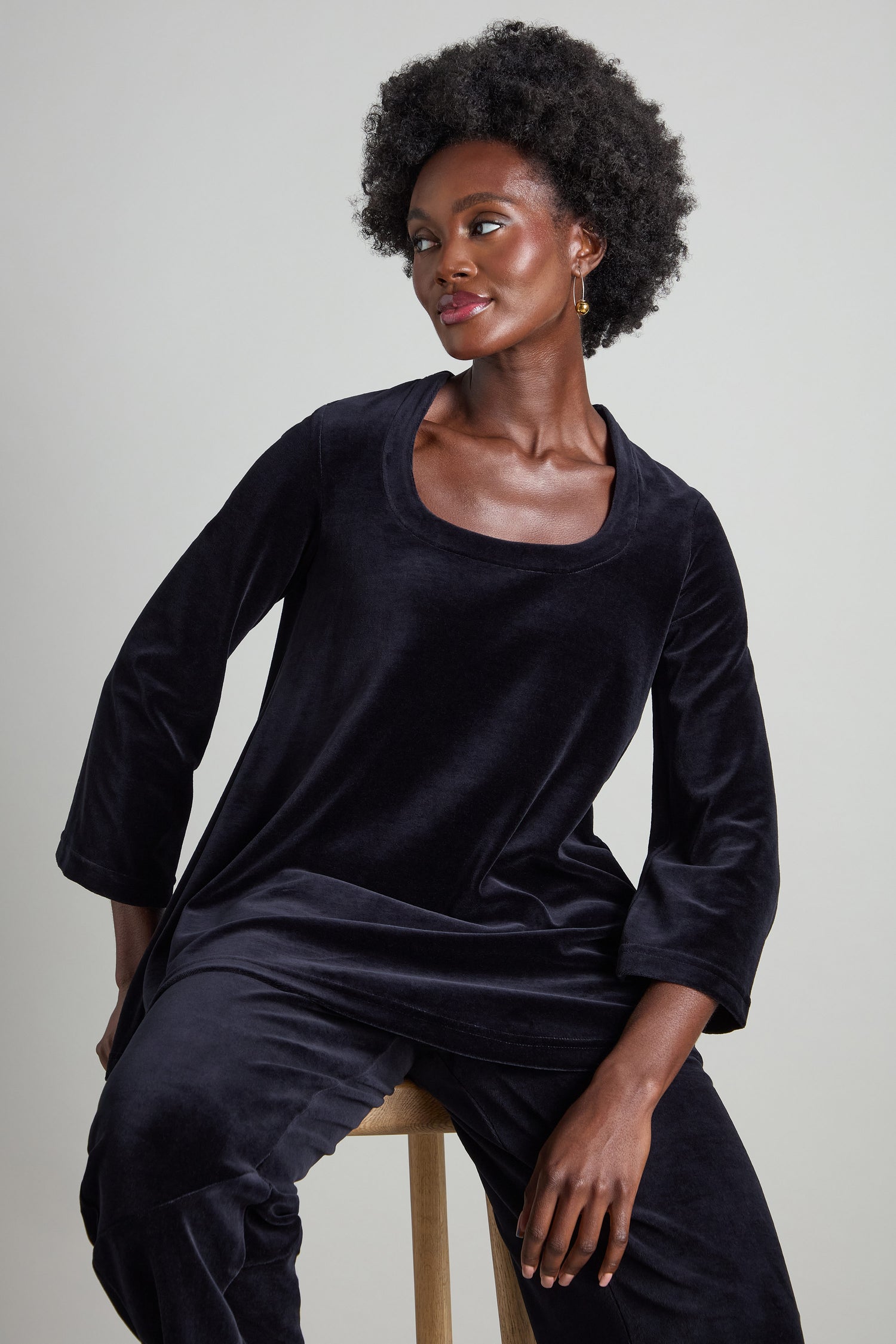 A woman sits gracefully on a stool, adorned in a Velvet Jersey Horseshoe Neck Top featuring elegant flared sleeves.