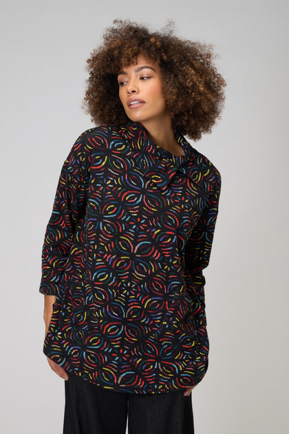 A person wears the Mosaic Jacquard Jersey Top—black, long-sleeved, with a colorful circular geometric pattern—while standing against a plain light gray background.