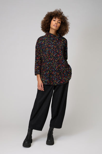 A person with curly hair wears the Mosaic Jacquard Jersey Top with a colorful circular pattern, relaxed black wide-leg pants, and black boots, standing against a plain light background.
