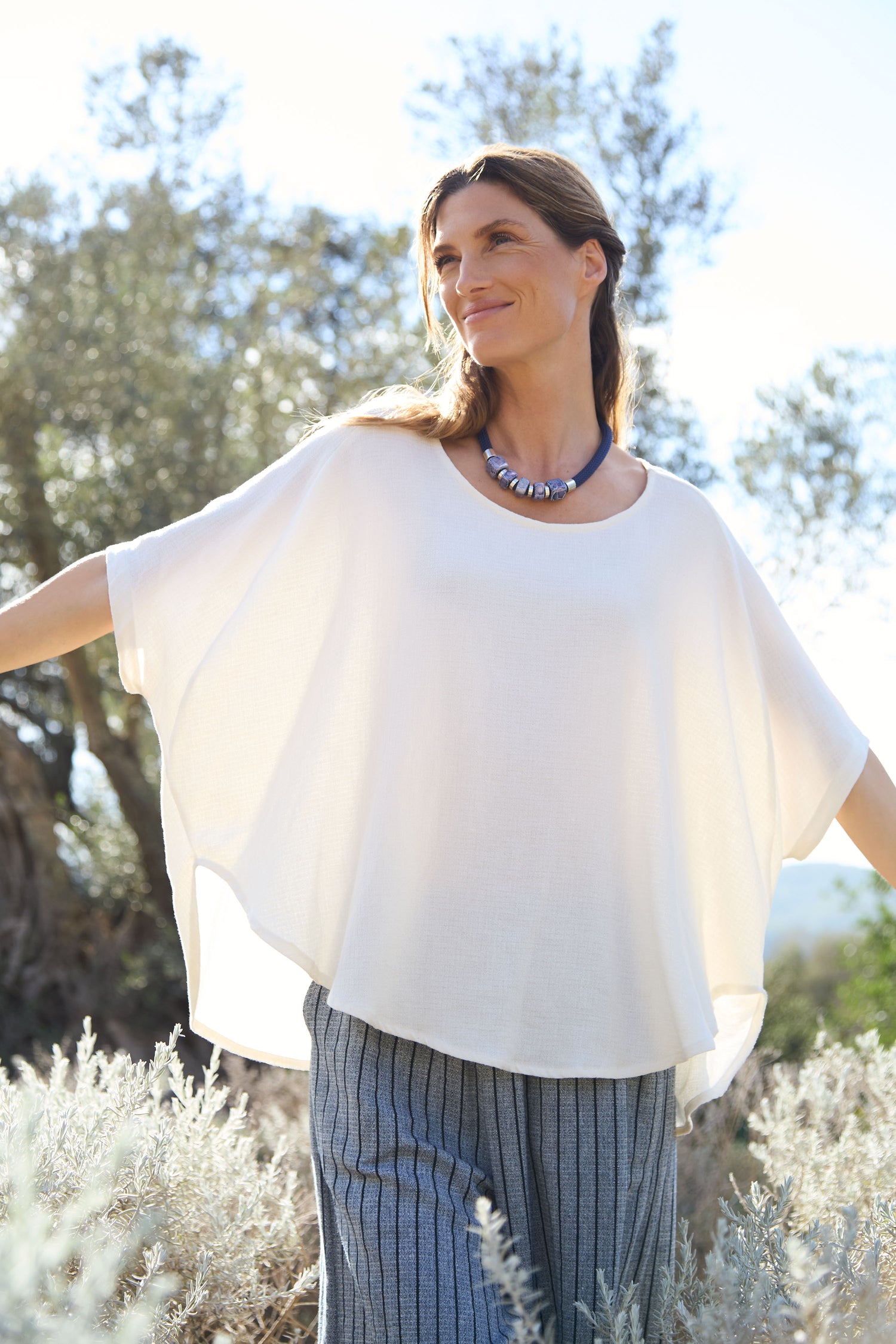 In the golden sunlight filtering through the trees, a woman gracefully extends her arms, wearing a white Linen Viscose Relaxed Top and striped pants. This serene scene stands as if it could be captured in an artistic photograph with code SPT6223-IHL marking its elegance.