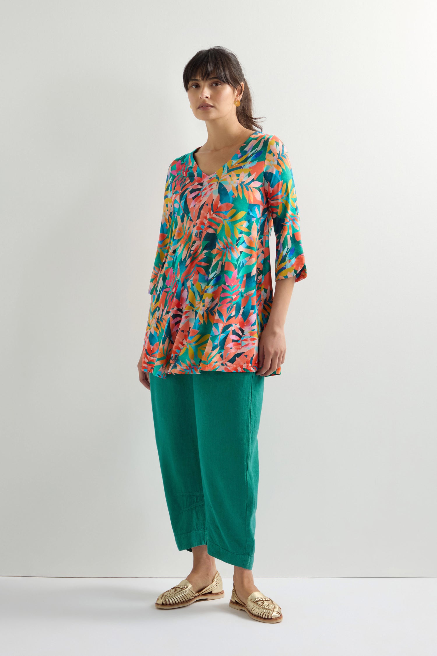 A person stands against a plain background wearing the Tropical Leaves Jersey Print Tunic, teal pants, and gold sandals.