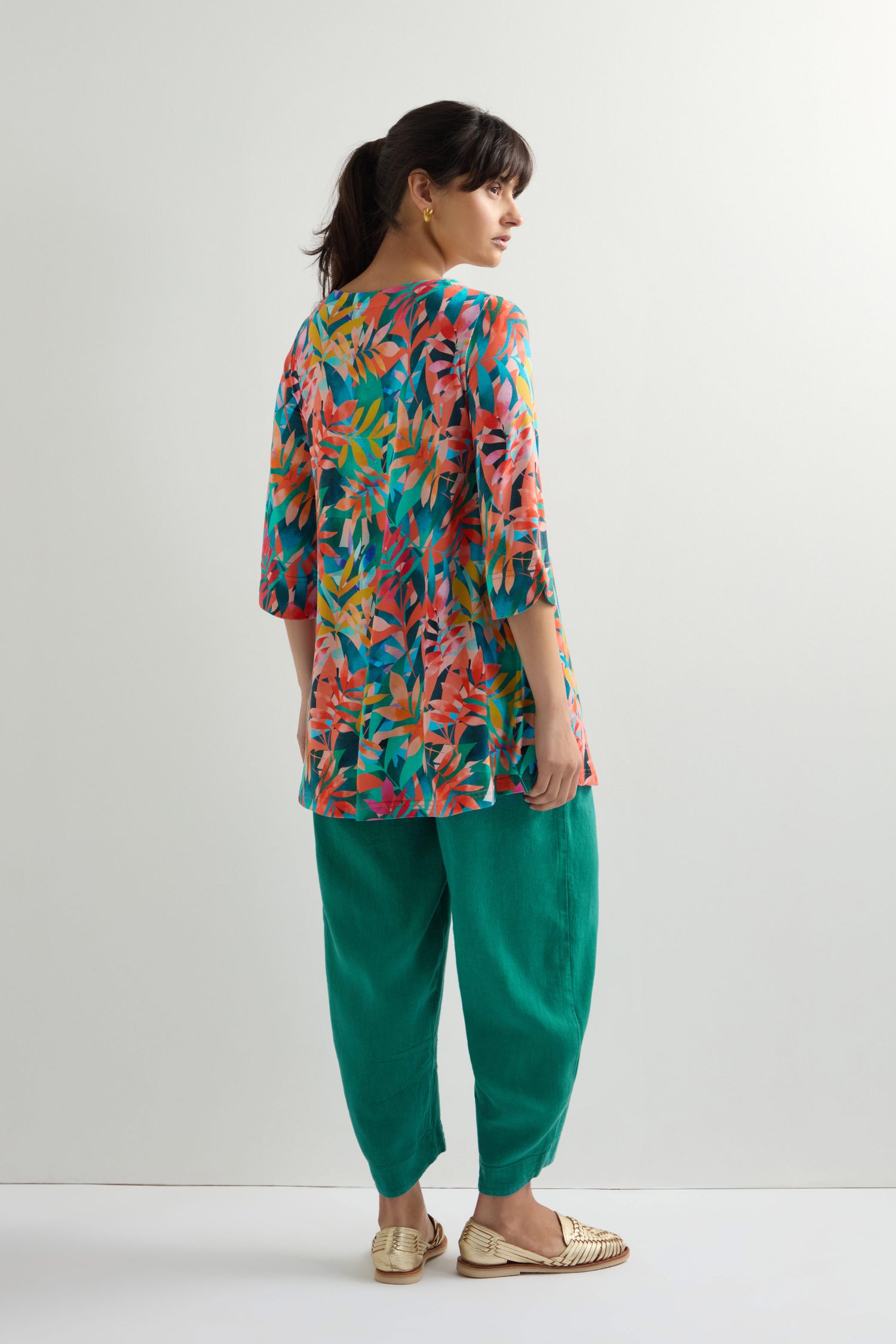 A person models the Tropical Leaves Jersey Print Tunic with green pants and beige shoes, set against a plain background.