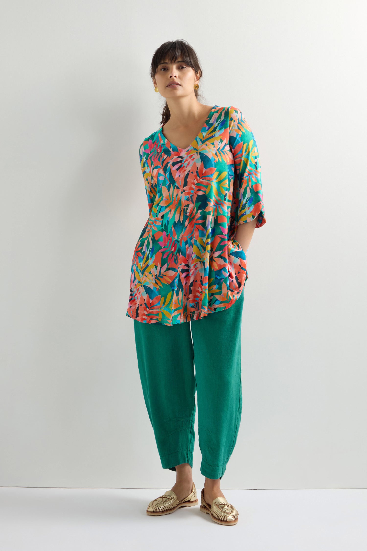 A person wearing a colorful Tropical Leaves Jersey Print Tunic, paired with green pants and gold shoes, stands against a plain white background.