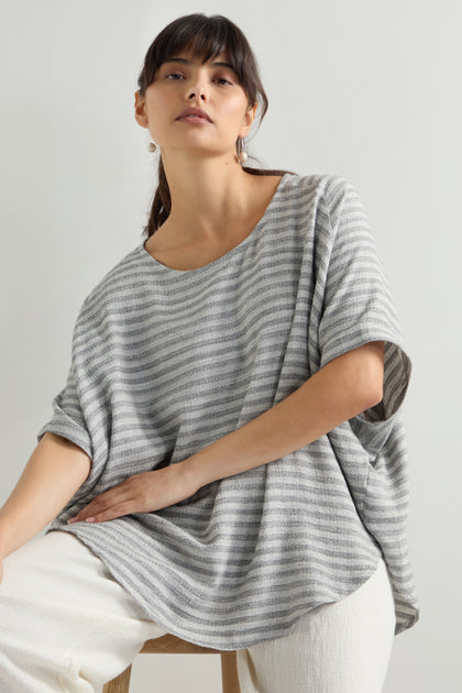 A person sits against a plain backdrop wearing the Textured Stripe Linen Viscose Top, characterized by its loose, oversized fit, and paired with white pants.