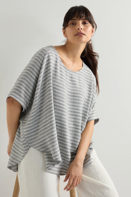 Wearing the Textured Stripe Linen Viscose Top, a person in gray and white stripes paired with cream pants sits on a stool against a plain background.