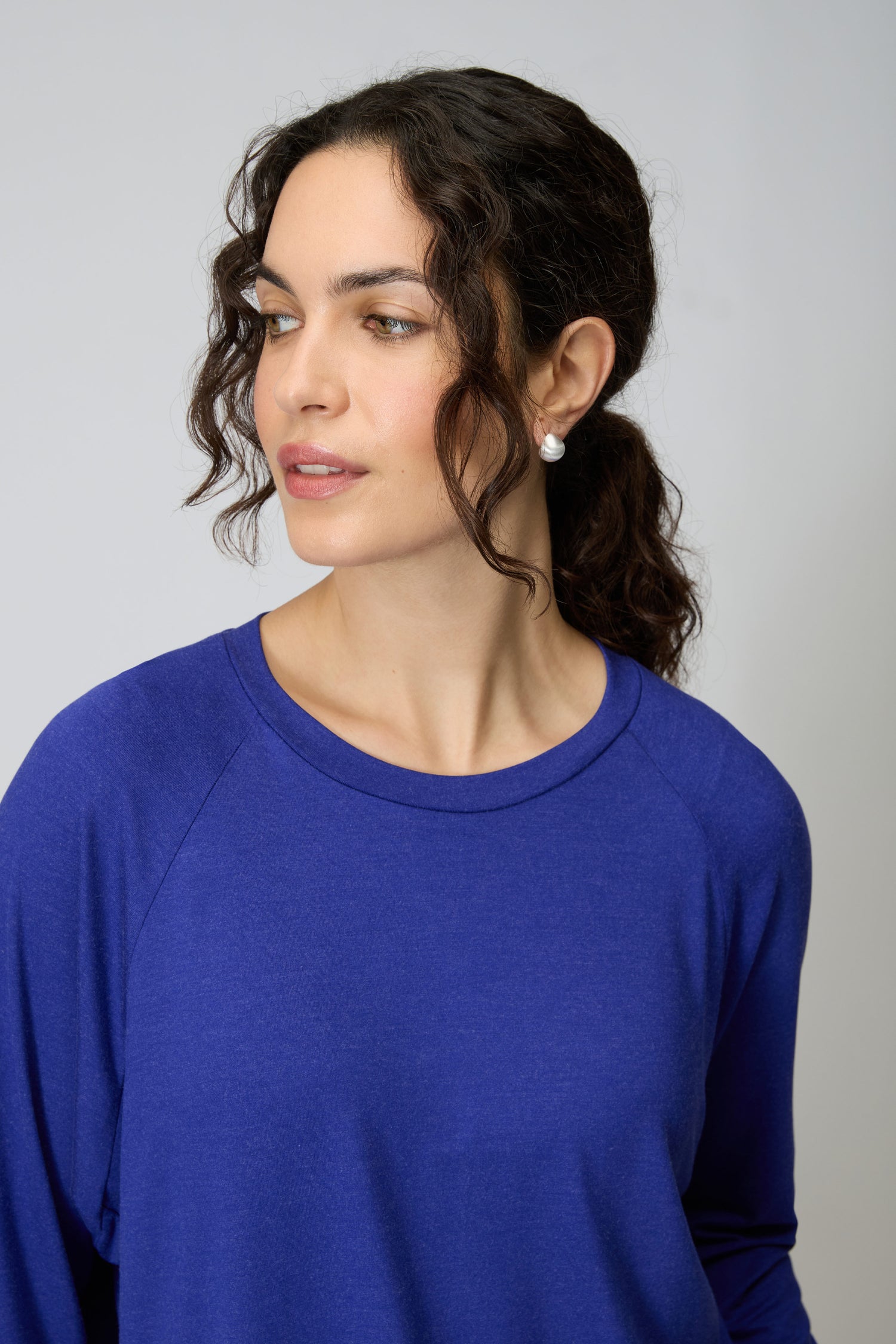Woman with wavy dark hair in a ponytail wears the Soft Viscose Jersey Boxy Top in blue and pearl earrings, looking slightly to the side against a plain background.