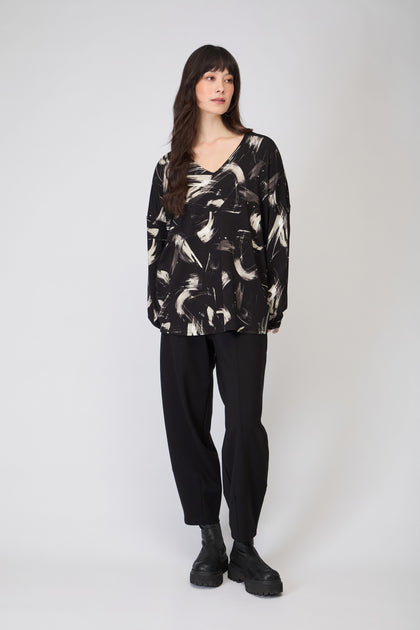 A woman stands facing slightly left, wearing the Painted Brushstrokes Jersey Top in black and white, paired with black pants and black boots against a plain light background.