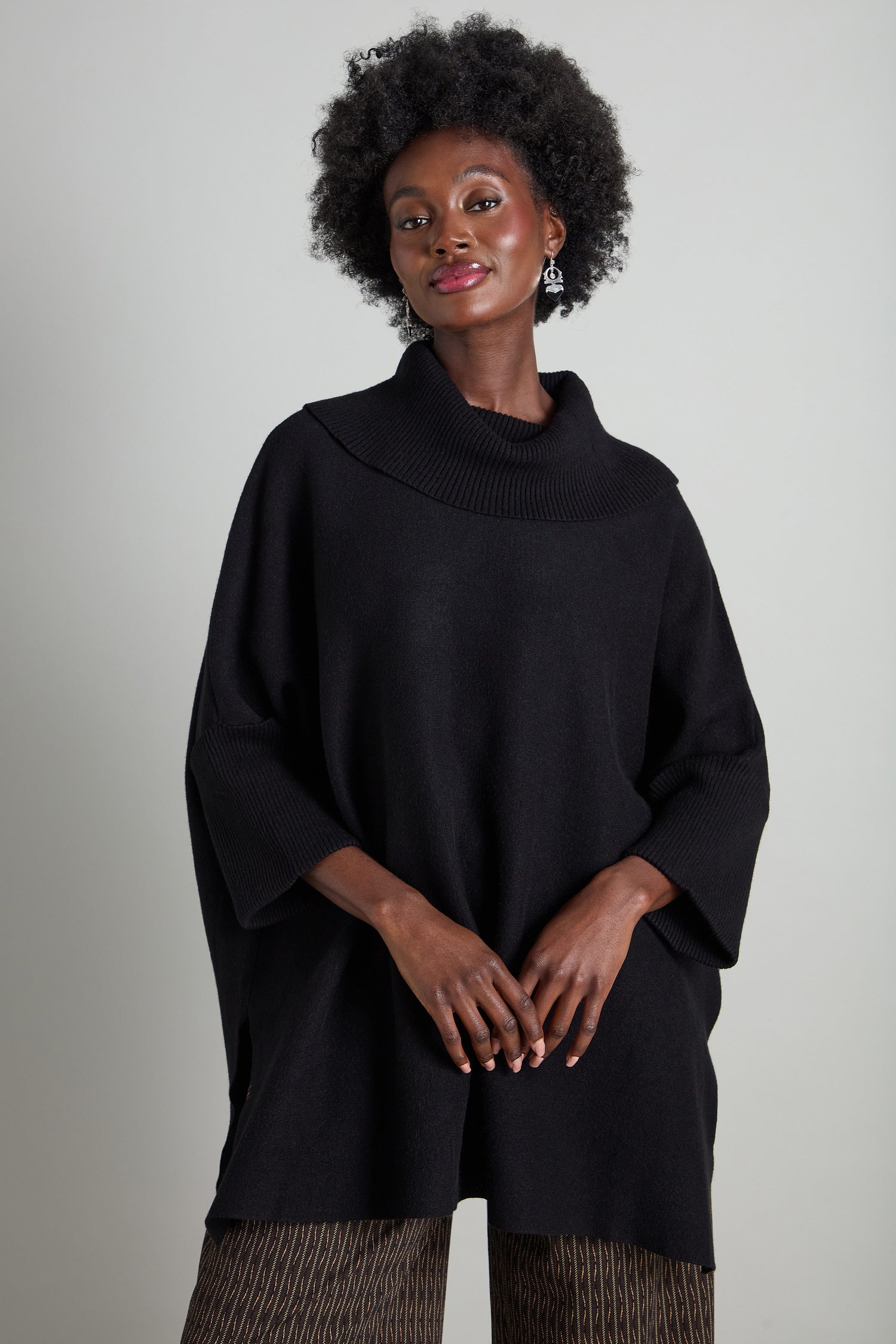 A person with curly hair wearing a Roll Neck Knit in black and patterned pants stands against a neutral background, smiling slightly.