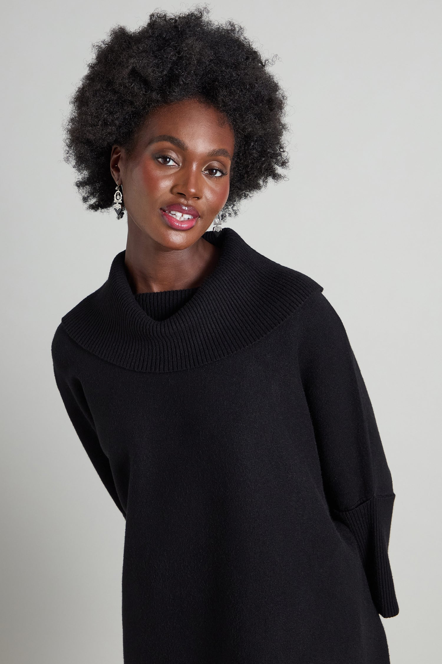 A person with curly hair wearing a Roll Neck Knit in black and earrings, standing against a neutral background.