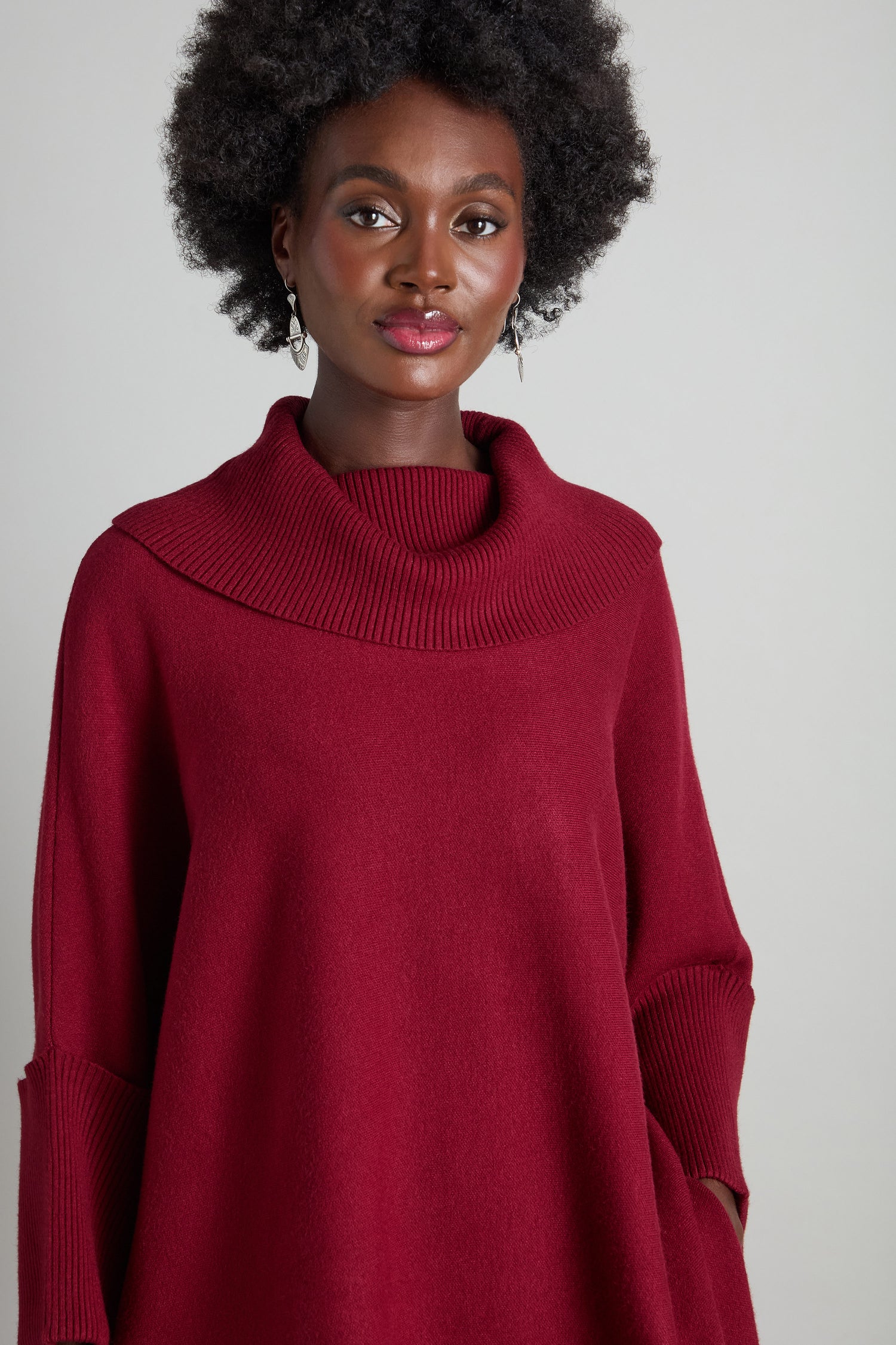 A woman stands against a plain background, elegantly dressed in the Roll Neck Knit, a red long-sleeve sweater that's ideal for layering.