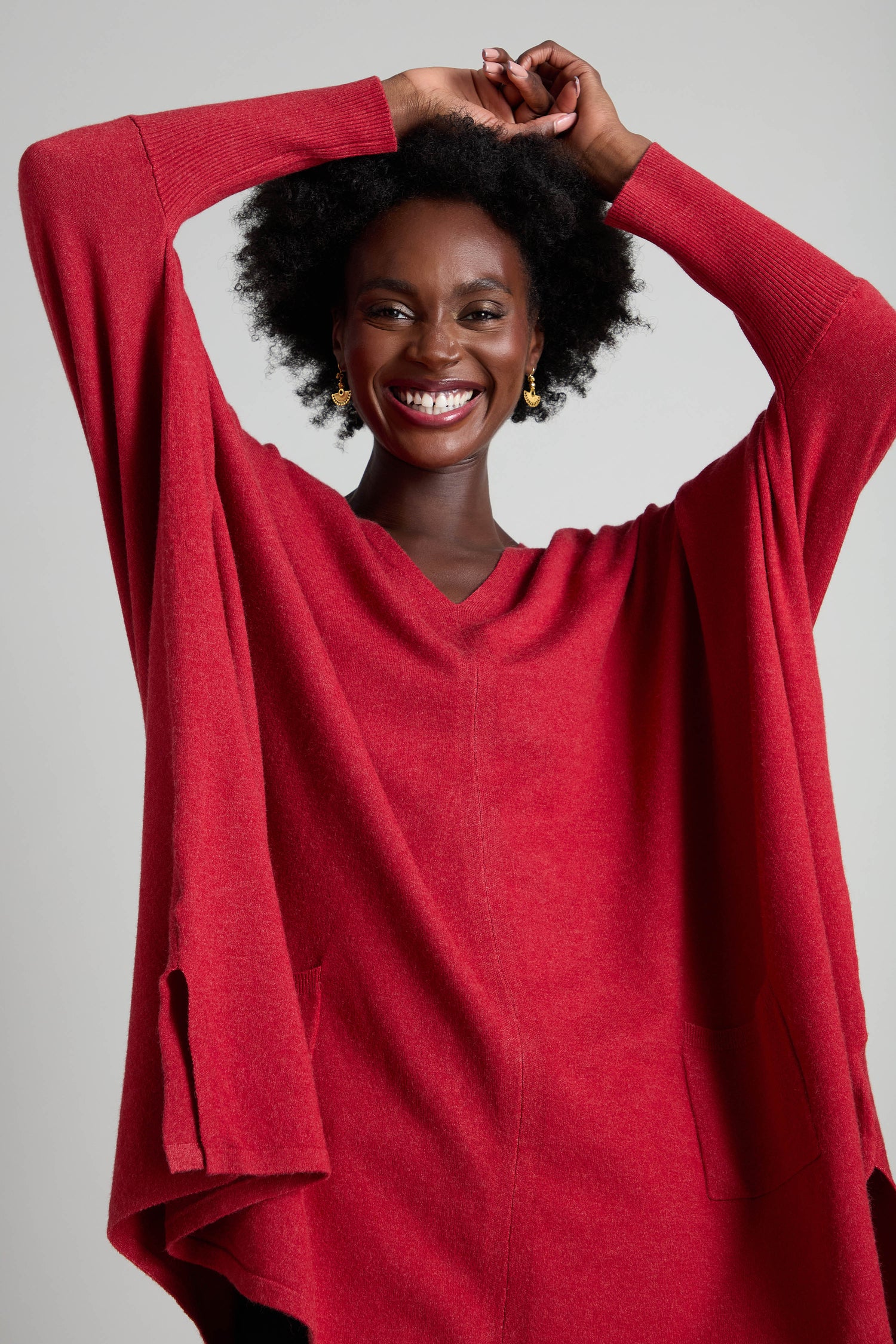 A person is smiling, adorned in a red V Neck Knit and gold earrings, with their hands raised above their head against a plain background. Ideal for layering during the cooler months, this ensemble is both stylish and cozy.