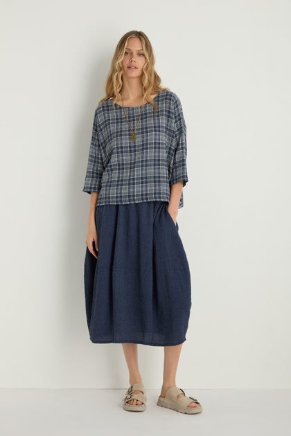 A woman stands against a plain white wall wearing a blue plaid top, the Crinkle Bubble Skirt in navy with an elasticated waist, beige sandals, and a gold necklace.
