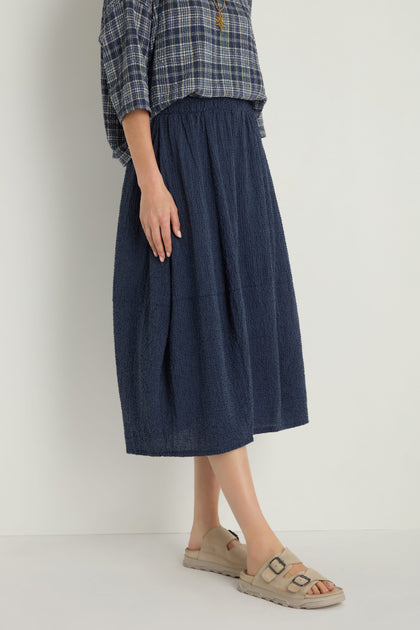 Someone wears a blue plaid shirt, beige sandals, and the Crinkle Bubble Skirt—a navy blue textured cotton-blend skirt with an elastic waist—while standing against a plain light background.