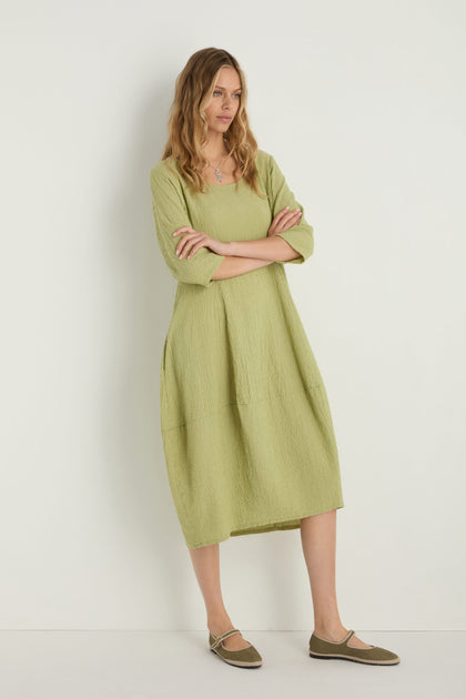 A woman stands against a plain white wall with arms crossed, wearing the Crinkle Bubble Dress in light green, a relaxed fit textured cotton blend, paired with matching green flat shoes.