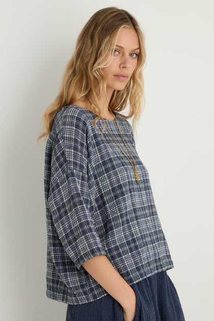 A woman with wavy blonde hair models the Check Boxy Top with a long necklace and a navy textured skirt, standing against a plain light background.