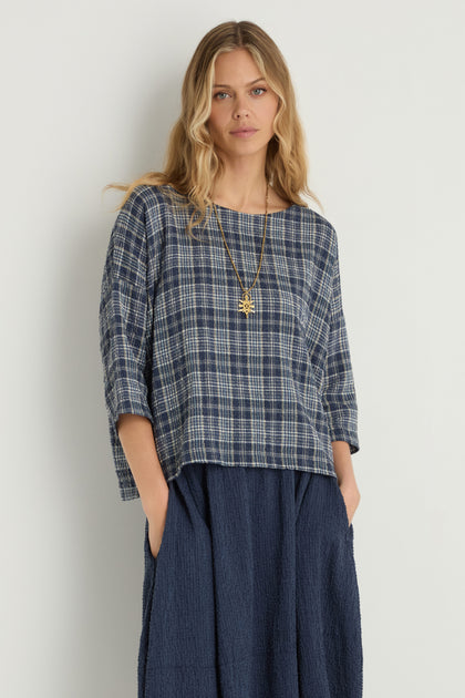 A woman with long blonde hair wears the Check Boxy Top in blue plaid and a navy skirt, accessorized with a gold necklace. The top is crafted from a soft textured cotton blend for comfort, set against a plain white background.