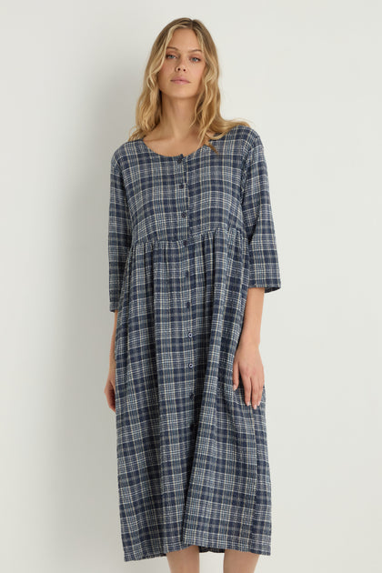 A woman stands against a plain background wearing the Check Button-Through Navy Dress, featuring a blue and white plaid Danish design with three-quarter sleeves.