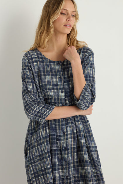 A woman stands against a plain white background wearing the Check Button-Through Navy Dress, looking to the side with one hand near her chin, reflecting timeless Danish design.