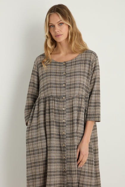 A woman with long blonde hair stands against a plain white background, wearing the Check Button-Through Taupe Dress featuring three-quarter sleeves and a relaxed gathered plaid design.
