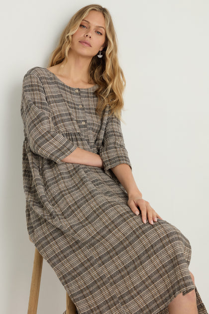 A woman sits on a stool wearing the Check Button-Through Taupe Dress, featuring long sleeves and chest buttons, against a plain light background.