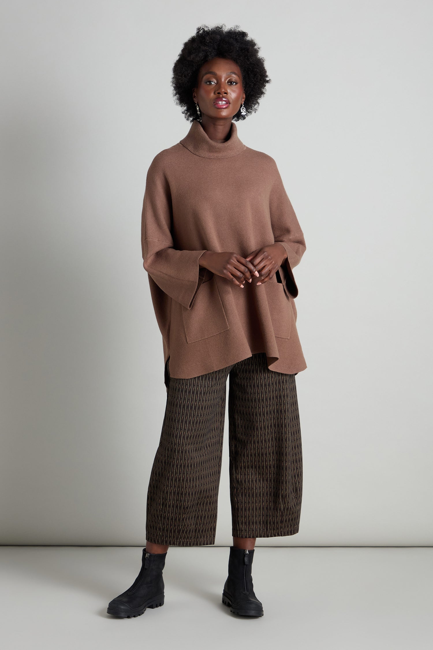 A person stands against a plain background, wearing the Roll Neck Pocket Knit in brown, paired with patterned wide-leg pants and black boots.