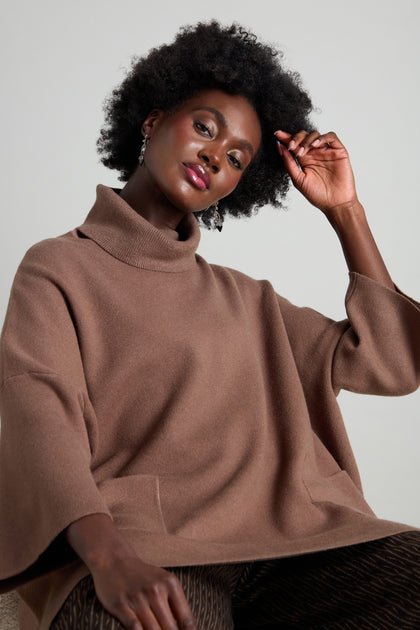 A person with curly hair is seated, donned in a Roll Neck Pocket Knit and patterned pants featuring oversized pockets, posing with one hand gently touching their hair.