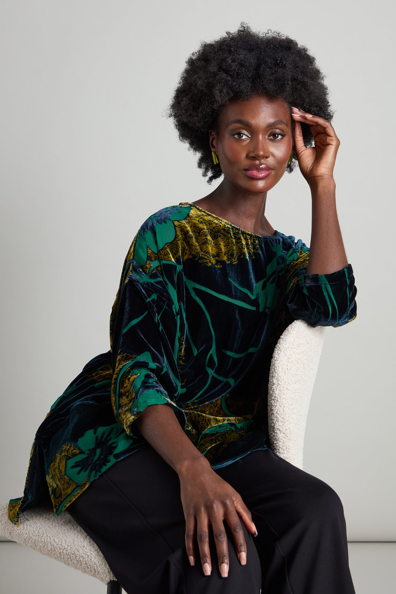 A person with an afro hairstyle sits stylishly on a chair, wearing the Lily Devore Tunic featuring green lily designs and matched with black pants, striking a pose with one hand resting on their head.