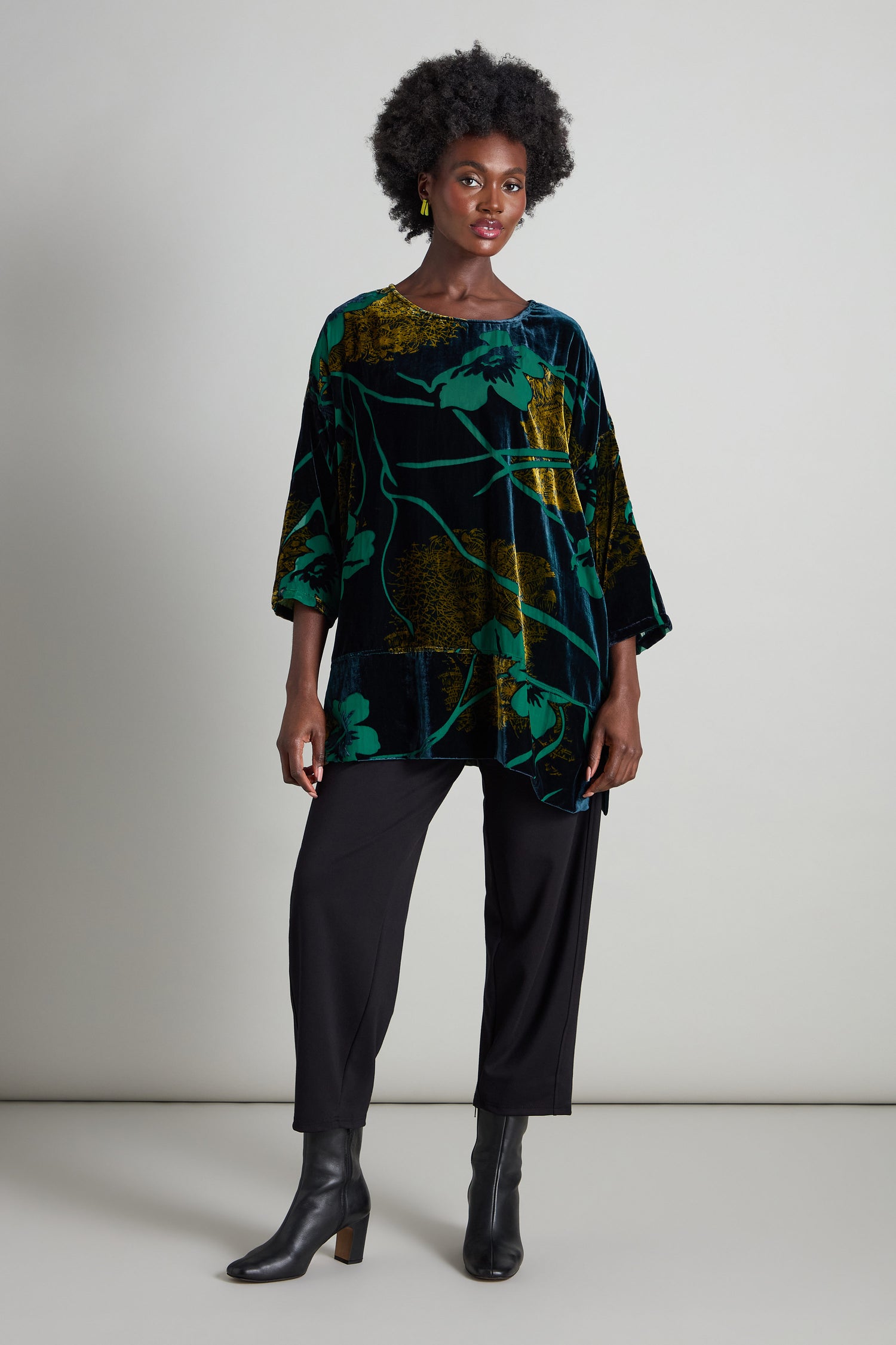 A person stands wearing the Lily Devore Tunic, its elegant green and black floral velvet pattern adorned with delicate green lily designs. Complemented by black pants and ankle boots against a simple backdrop, the outfit radiates sophistication.