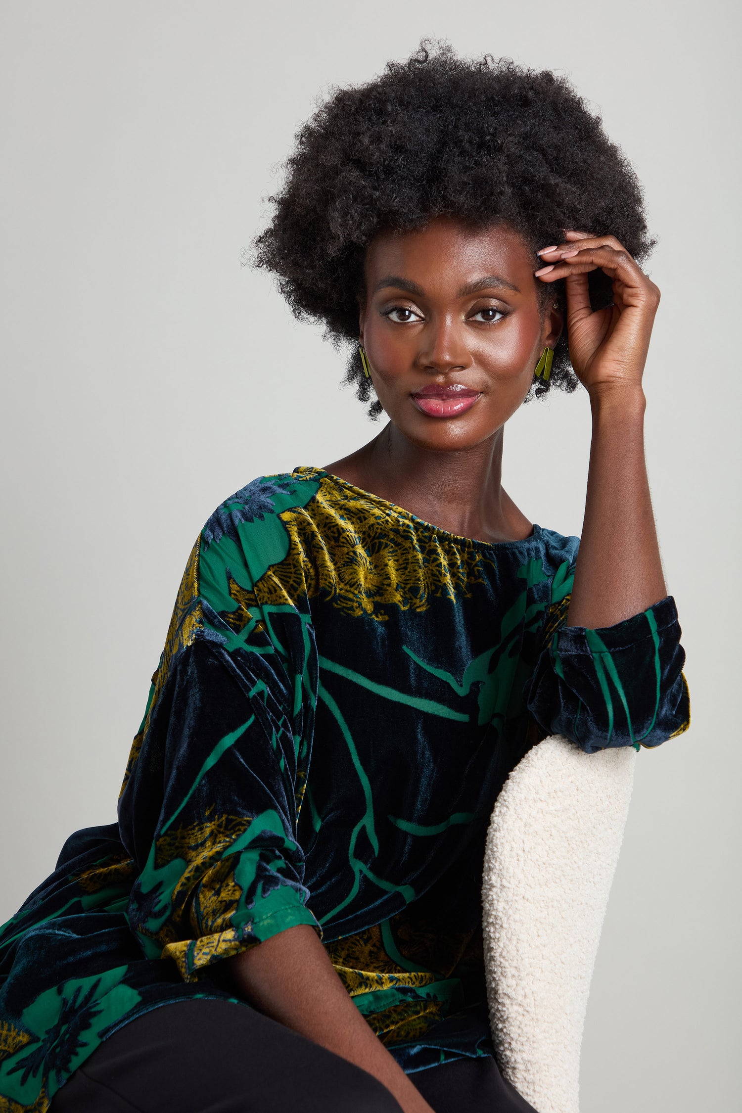 A person sits on a chair, wearing the Lily Devore Tunic with a green and black pattern adorned with delicate green lily motifs, smiling at the camera.