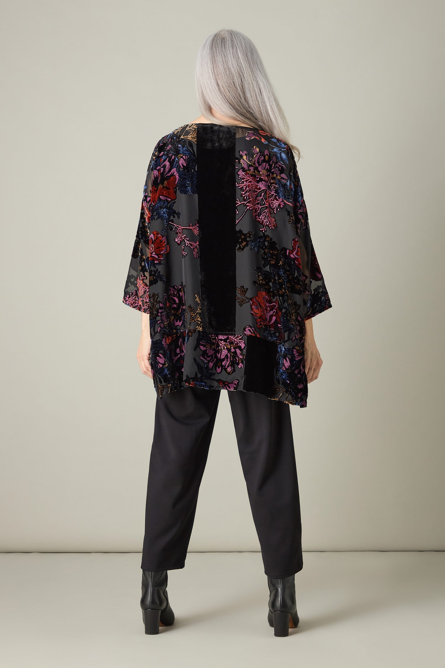 A person with long gray hair stands facing away, wearing a Cross Detail Devore Tunic adorned with floral patterns, alongside black pants and boots, against a plain background.