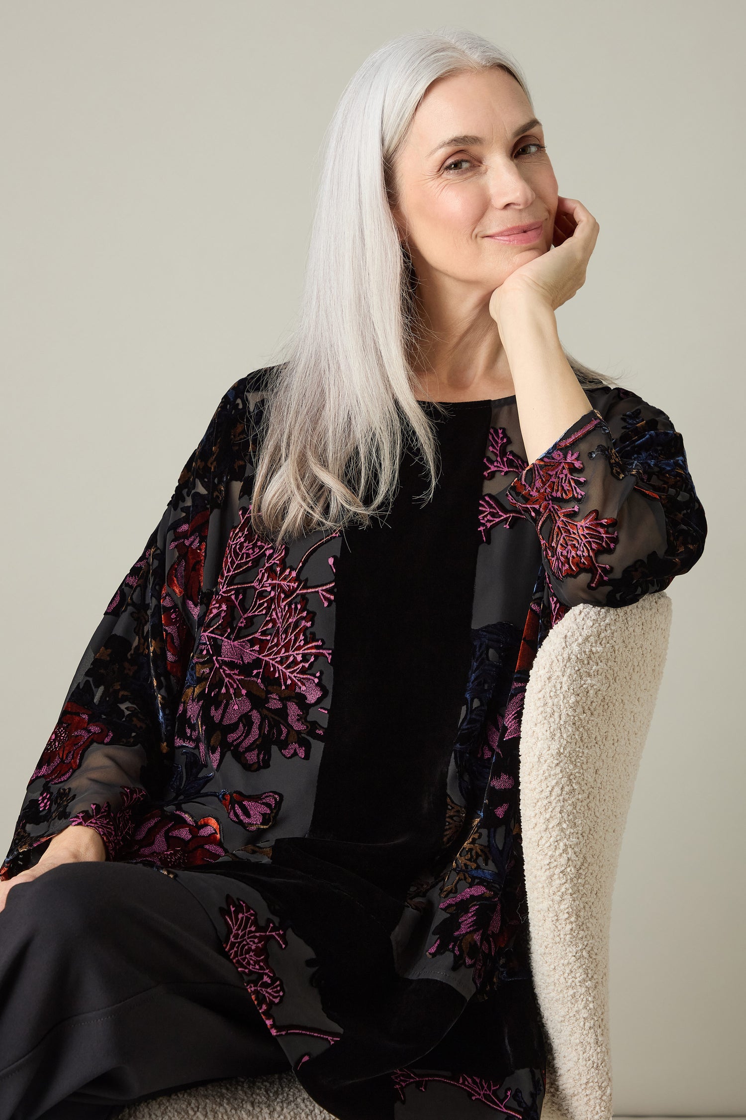 A woman with long gray hair sits on a chair, wearing the Cross Detail Devore Tunic, featuring floral patterns reminiscent of velvet devoré. She rests her chin on her hand and smiles slightly.