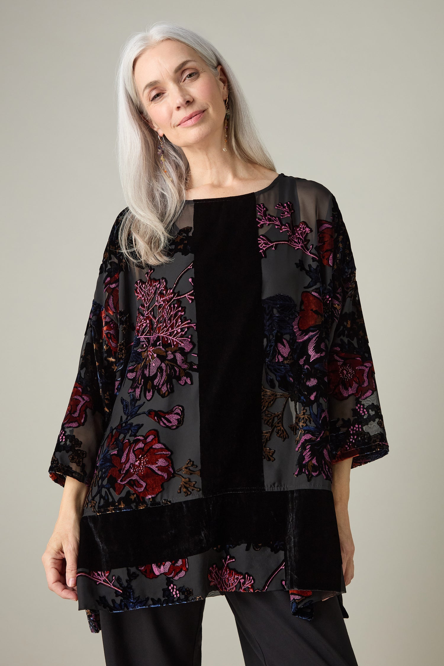 A person with long gray hair, wearing the Cross Detail Devore Tunic in black, stands against a neutral background, looking forward with a slight smile.