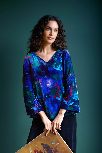 A woman with curly hair is wearing the Late Night Flora Velvet Top, showcasing a painting against a dark green background.