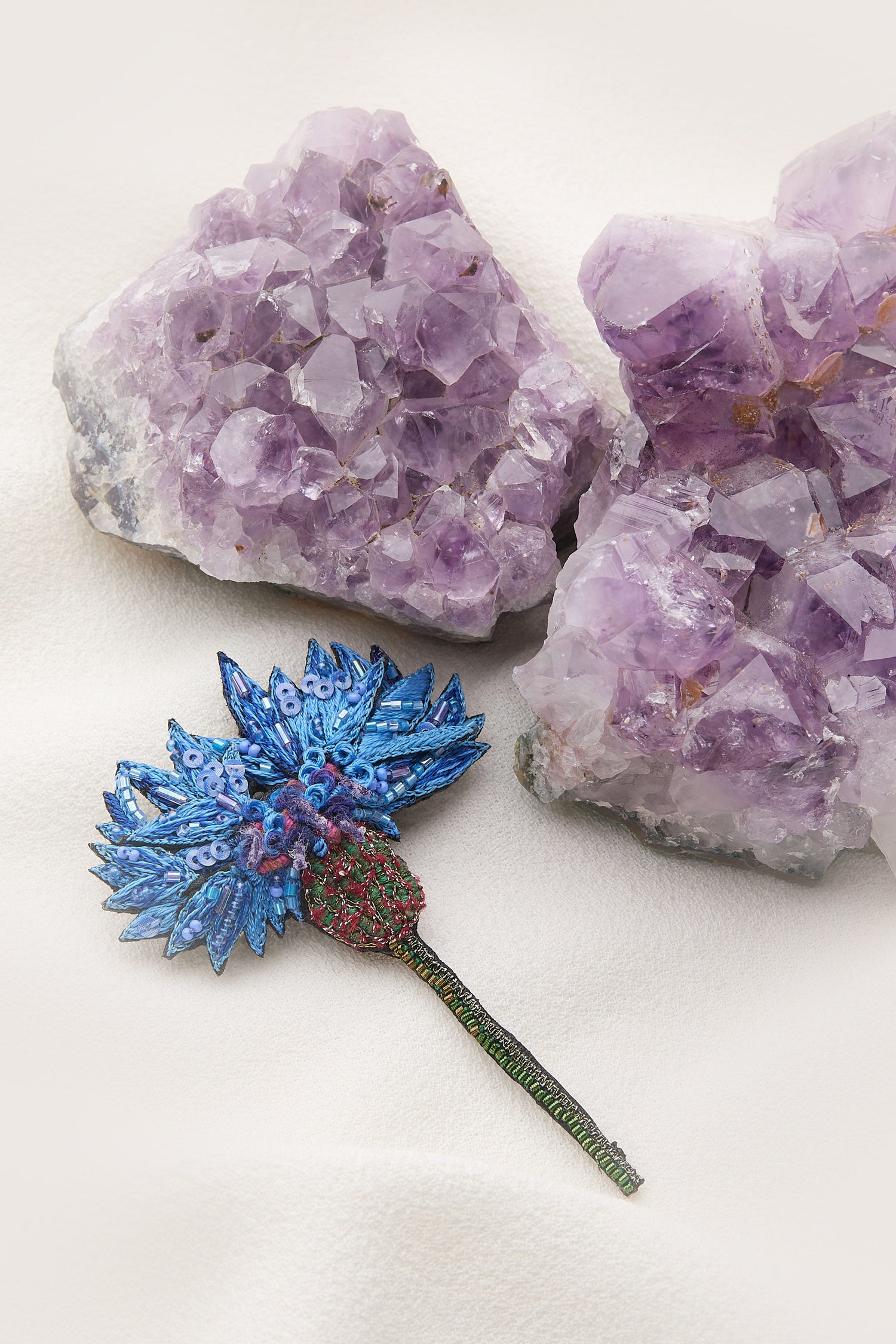 A Hand Embroidered Blue Cornflower Brooch, decorated with blue and green jewels, lies on a white surface beside two large purple crystal clusters, shimmering with the allure of intricately embroidered details.