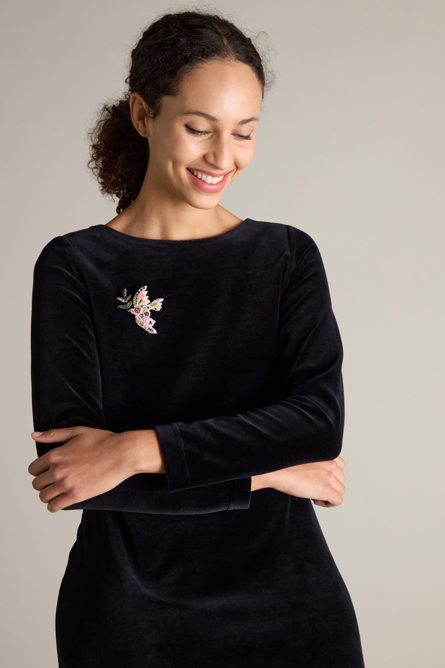 A woman with curly hair smiles, looking down with arms crossed. She is wearing a long-sleeved, dark-colored dress adorned with a Trovelore Hand Embroidered Peace Dove Brooch on the left side of the chest.