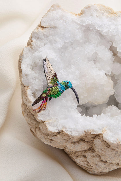 A detailed, colorful Hand Embroidered Sequin Hummingbird Brooch is placed on a large open geode with crystals inside, set against a light fabric background.