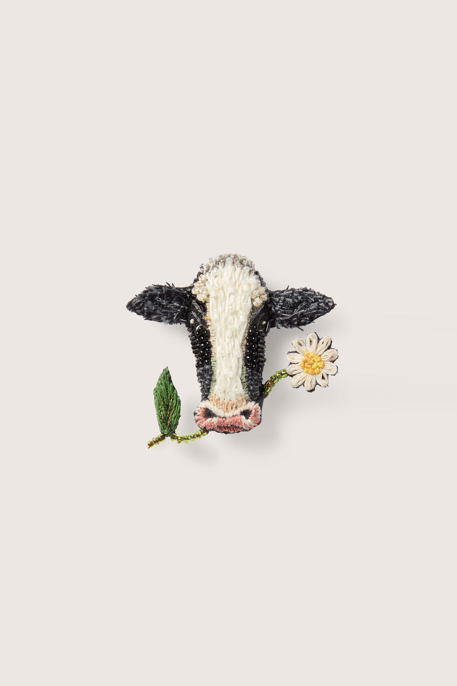 The Hand Embroidered Cow Brooch features a black and white patterned face holding a daisy in its mouth. The plain light-colored background accentuates the whimsical touch with intricate sequin and beaded detailing.