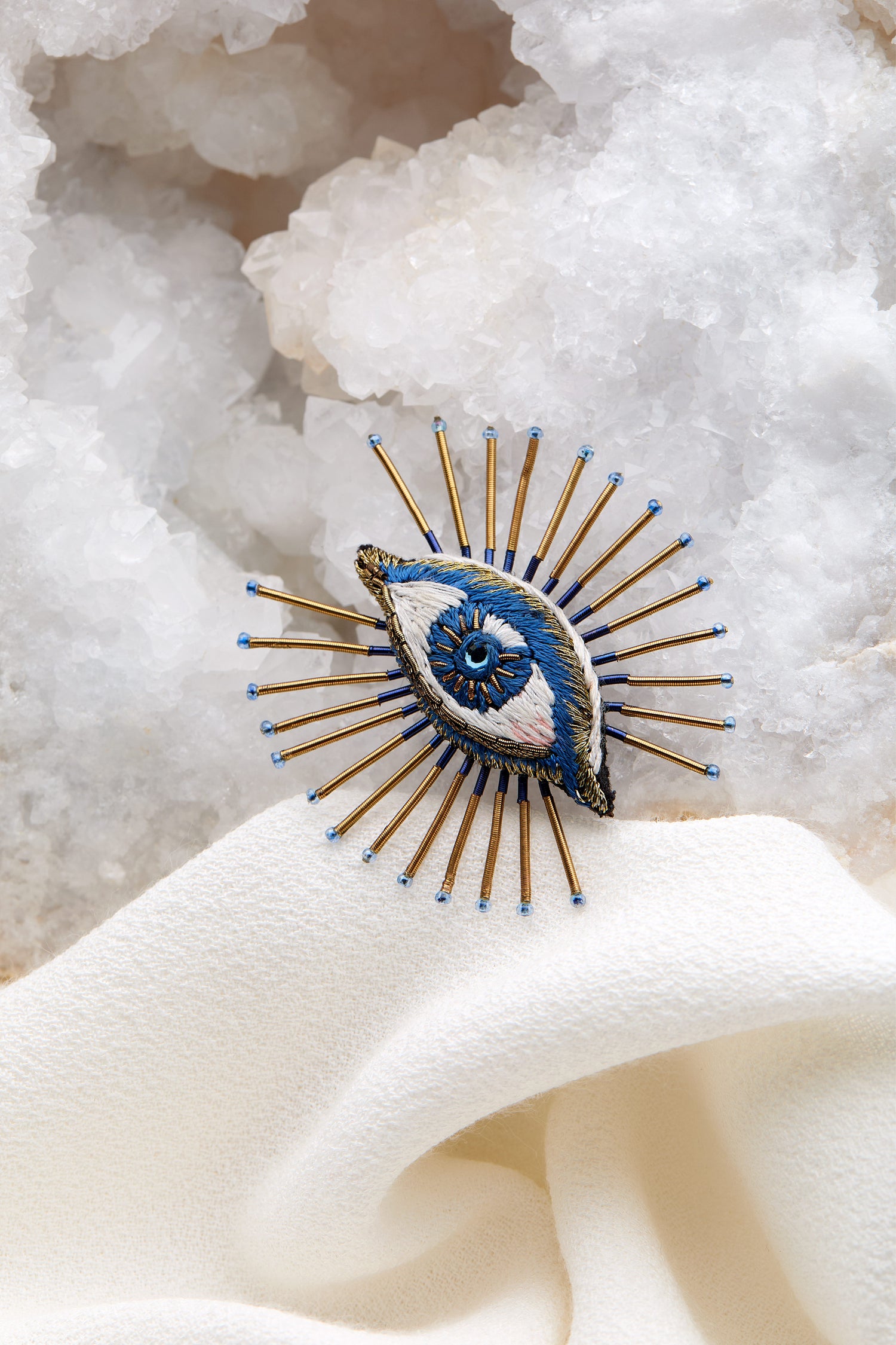 The Hand Embroidered Sea Mystic Eye Brooch, adorned with intricate craftsmanship, rests gracefully on a white cloth amid a crystalline background, its design mirroring an eye framed by fine metal pins.