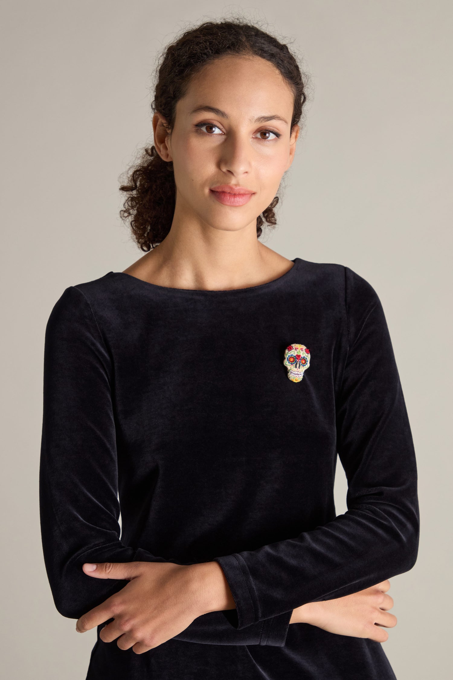 A woman with curly hair, wearing a black long-sleeve top adorned with beaded embellishments and a Hand Embroidered Calavera Brooch, stands with her arms crossed.