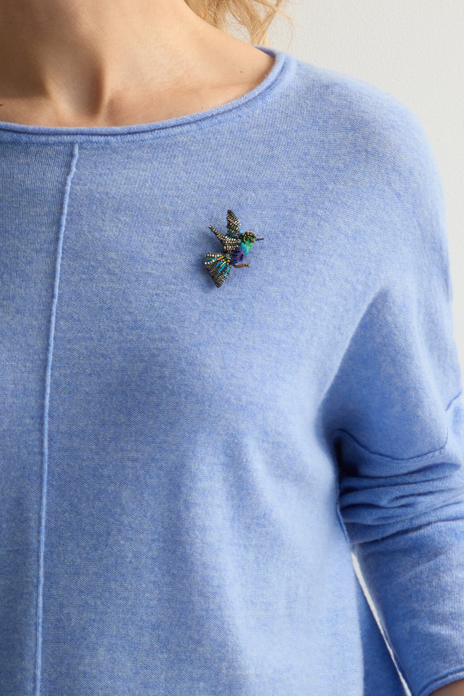 A person in a light blue sweater is adorned with a Hand Beaded Crowned Woodnymph Brooch from Trovelore Jewelry, gracefully perched on their shoulder.