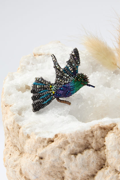 The Hand Beaded Crowned Woodnymph Brooch, reminiscent of Trovelore Jewelry, showcases exquisite black, green, blue, and purple beads. It elegantly sits on a textured white and beige stone surface.