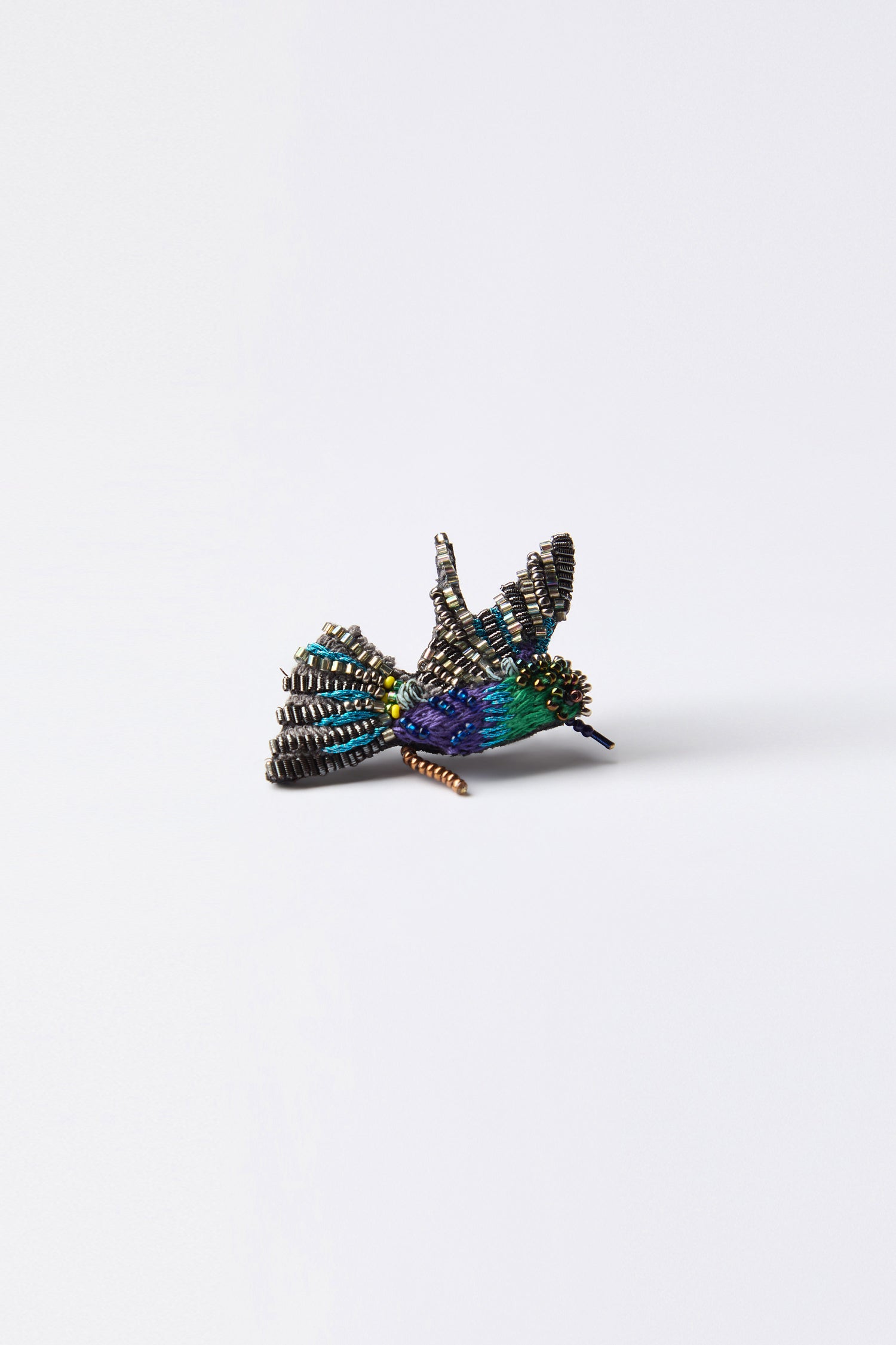 Against a white background, the Hand Beaded Crowned Woodnymph Brooch, a vibrant bird sculpture by Trovelore Jewelry, showcases exquisite artistry.