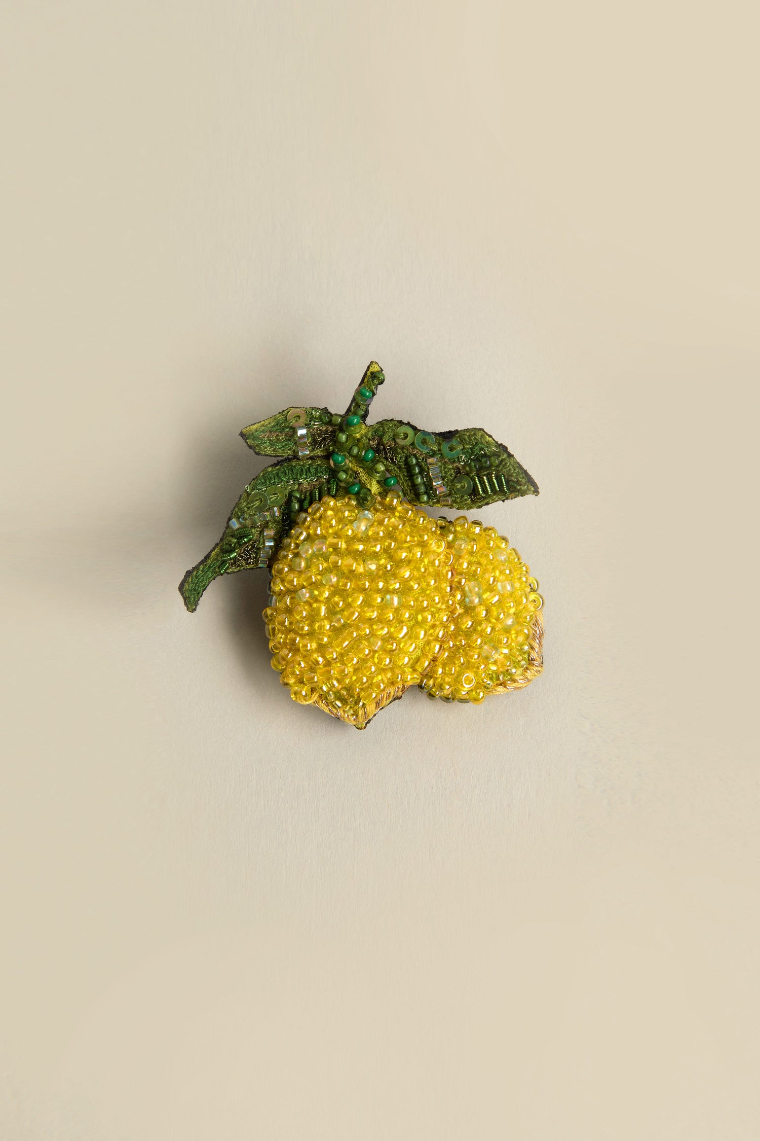 Discover Mediterranean beauty with our Hand Beaded Amalfi Lemons Brooch: vibrant beaded lemons and lush green leaves on a soft beige background, capturing sun-kissed elegance in its intricate design.