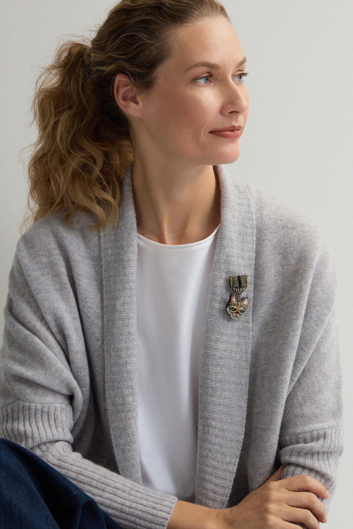 A woman with curly hair in a ponytail wears a gray cardigan over a white shirt, adorned with a Hand Embroidered Pussy Cat Brooch, looking to the side against a plain background.